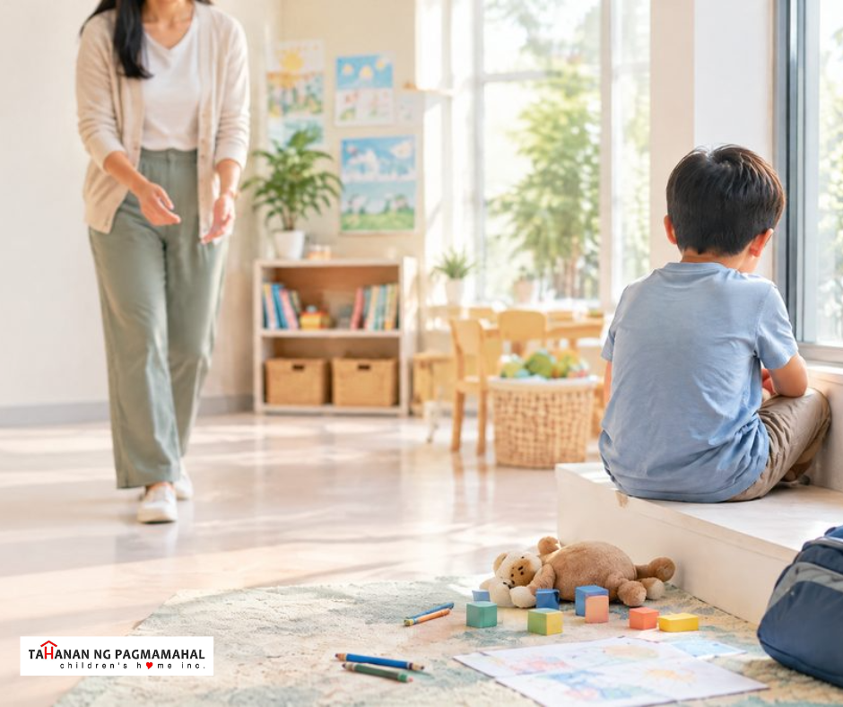 Filipino child sitting quietly by a bright window as a caring adult approaches in a community room, with unused toys and school supplies nearby.